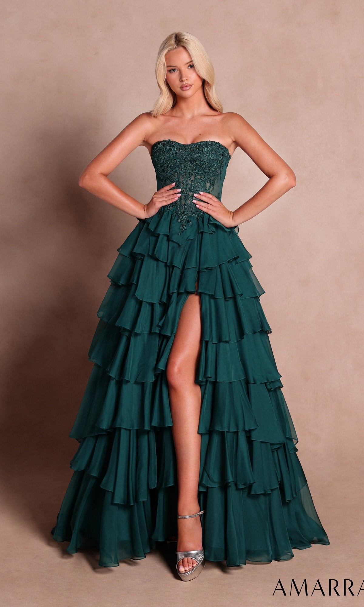 A woman stands against a plain beige background wearing the Amarra 89687 long prom dress in emerald—a strapless gown featuring ruffled details and a high front slit—paired with silver high heels.